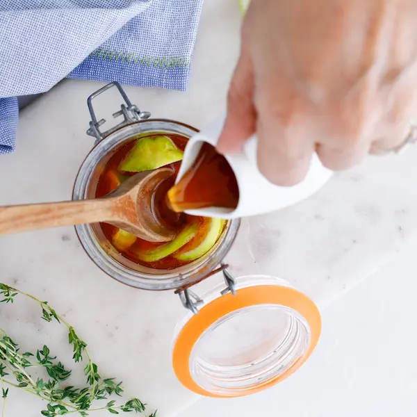 a jar of honey and lemons, with a hand pouring some thyme and ginger tea into the honey for a homemade cough syrup